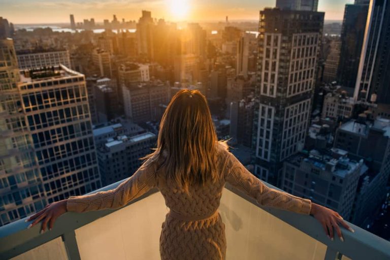A women stands on the balcony of a penthouse that over looks a city as the sun sets.