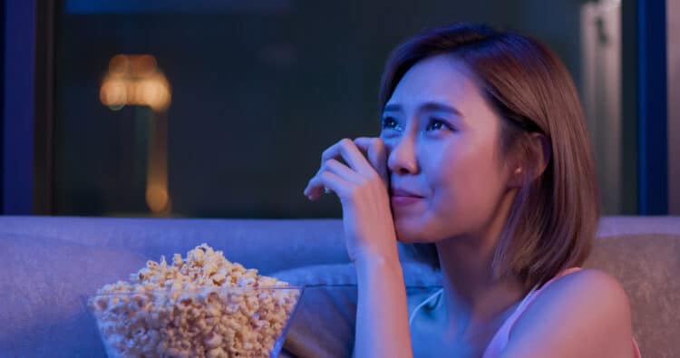 A young woman eating popcorn cries while sitting on a couch watching a movie after her breakup.