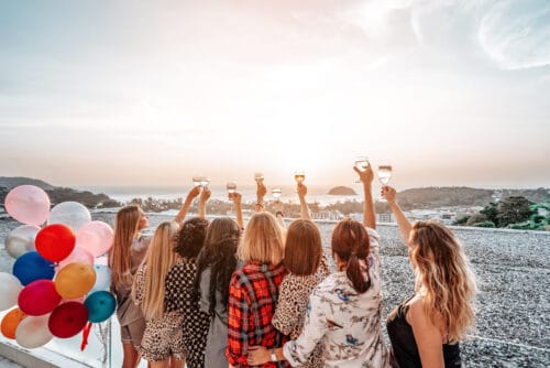Women at a divorce party toast the sunset from a balcony decorated with balloons.