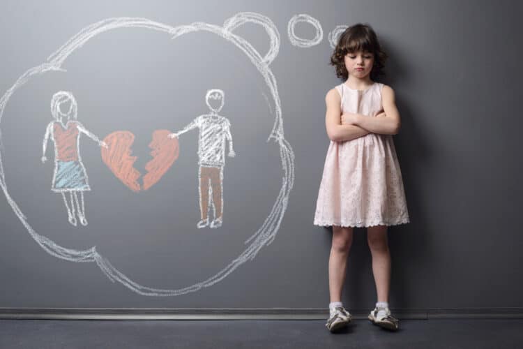 Little girl crossed her hands and looks very unhappy. Depicted divorce of the parents on the grey wall with chalk